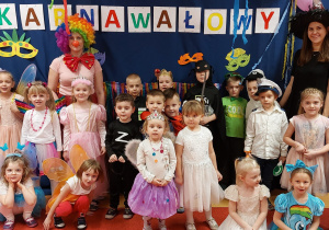 Zdjęcie przedstawia grupę dzieci wraz z paniami. Wszyscy stoją na tle dekoracji karnawałowej. Każdy ma na sobie kolorowy strój.